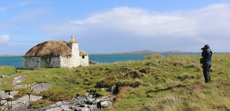 A person standing nearby a thatched rural cottage with the sea in the background