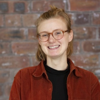 A headshot of Monica smiling, wearing red glasses