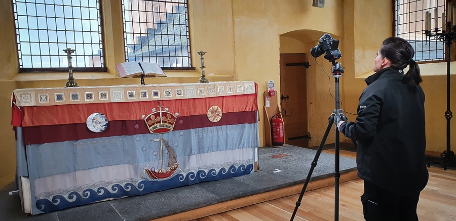 A person with a camera and tripod facing a large, historic tapestry