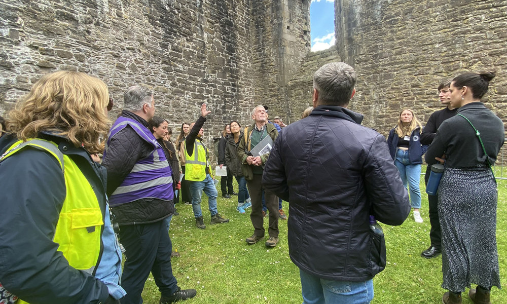 A group of people, some wearing high vis vests, standing on the grass beside a historic stone building, watching someone as they give them a talk