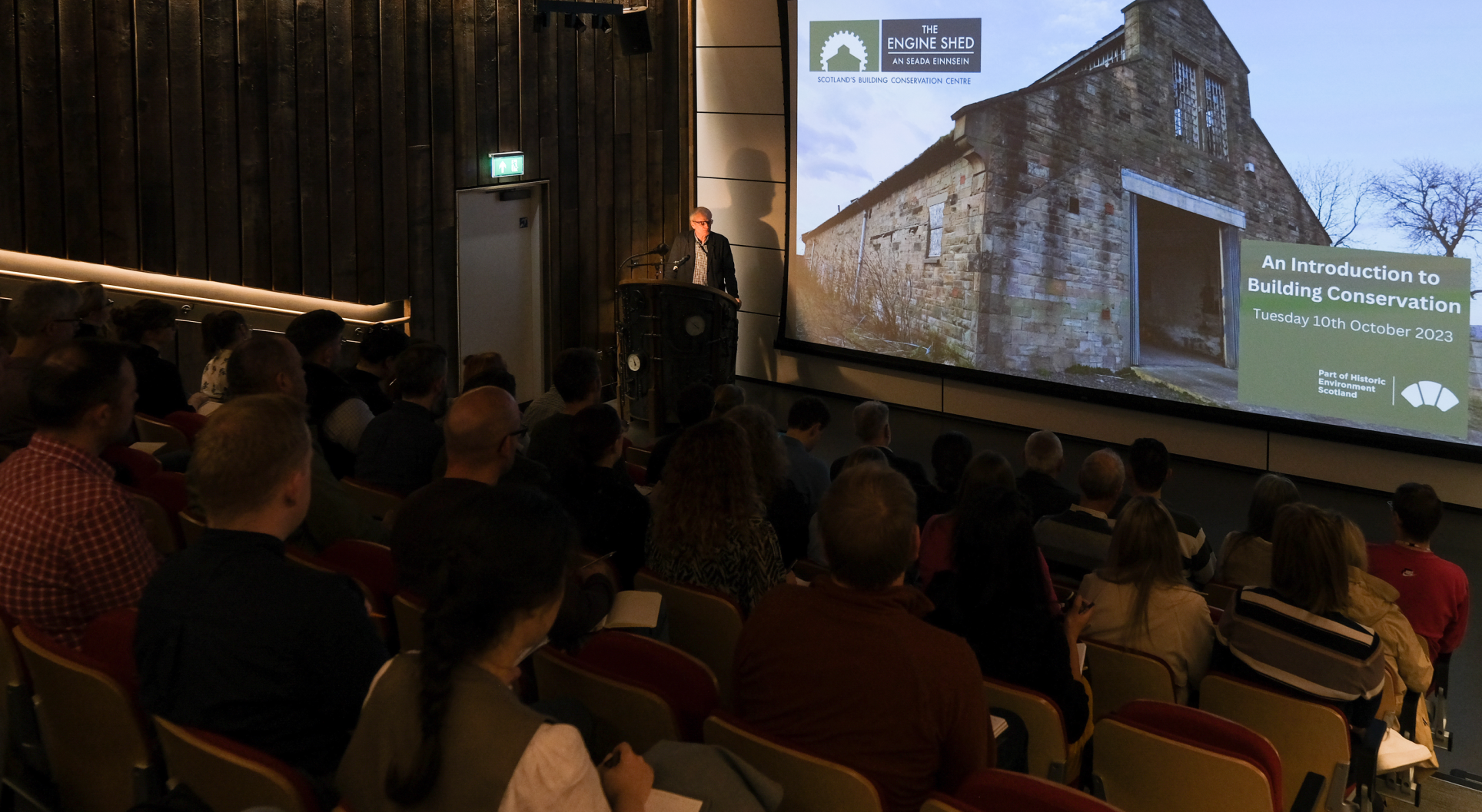 A dark auditorium at the Engine Shed is filled with people looking down at a large presentation screen and a speaker standing on the left corner of the image. 