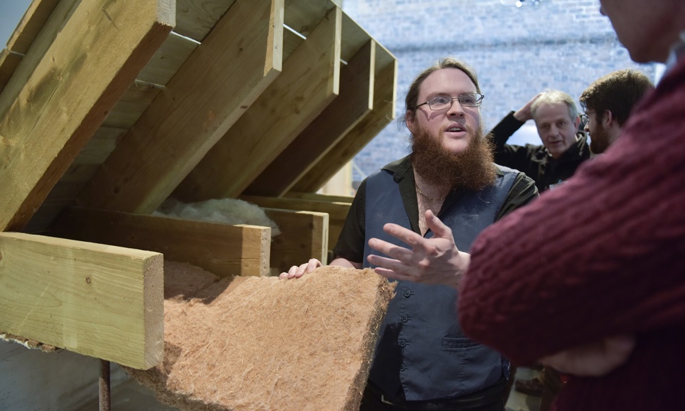 People are in conversation next to a model of the interior of the eaves of a roof. A piece of rough, flexible, beige insulation material is being shown as part of the conversation.